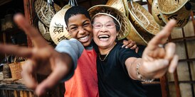 A teenage boy and an older woman smile as they extend their arms and give the peace sign with their hands while standing in front of a display of straw hats.