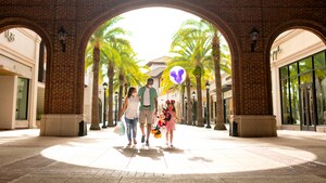 Wearing face masks, a mom, dad and their daughter, who is holding a large Minnie plush and a Mickey balloon, walk under an arch at Disney Springs