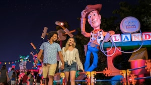 Una joven pareja sonríe mientras camina cerca de la entrada de Toy Story Land por la noche