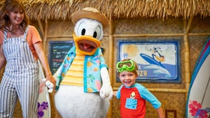 Donald Duck walks hand in hand with a woman and her son in front of a restaurant styled like a hut 