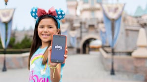 A young girl wearing Mickey Ears holds up a smartphone with the Play Disney Parks app open