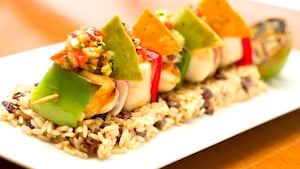 A skewer with grilled meat and vegetables topped with salsa and 4 nacho chips, atop rice and beans
