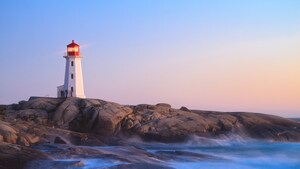 A lighthouse stands above smooth rocks and ocean waves at twilight
