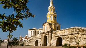 Una pared de piedra con tres puertas separa una plaza de una ciudad con edificios modernos y coloniales