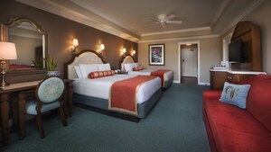 A wooden desk with padded chair sits next to two matching beds with a TV mounted on the opposite wall