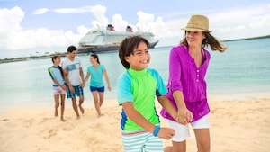 A woman dressed in beach attire smiles while walking hand in hand with her son on a sandy beach in front of a Disney cruise ship