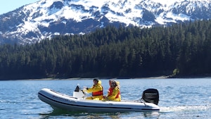 Um grupo de pessoas em um bote motorizado navegando pelas águas perto de um litoral arborizado, contra o pano de fundo de uma montanha com neve