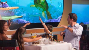 A family of 4 sit at a dinner table and wave to Crush the sea turtle who swims by on a large screen