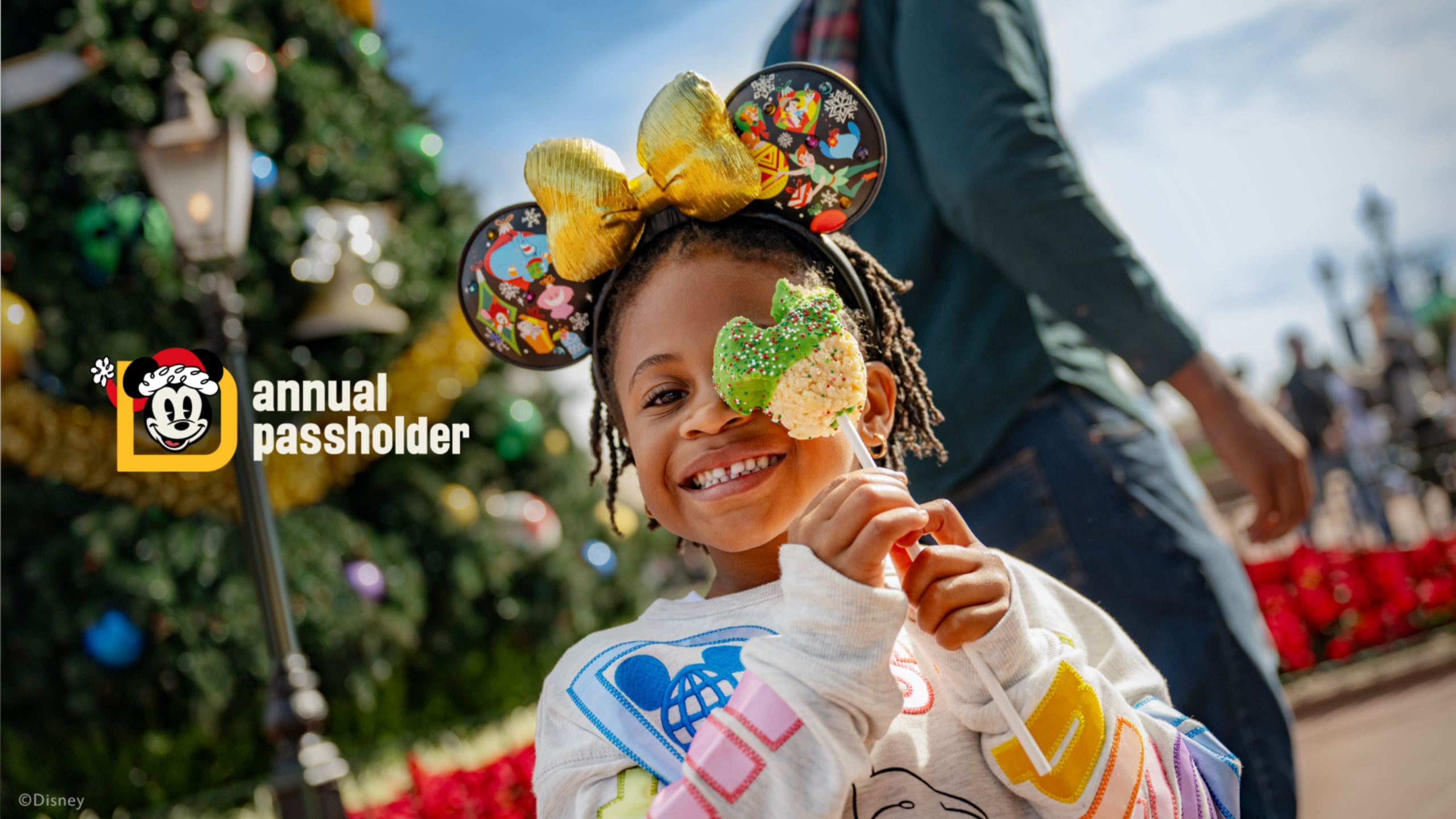 A young girl wearing a Christmas themed Minnie Mouse ear headband and holding a crisped rice treat with text that says ‘Annual Passholder’ next to a D with Santa Mickey Mouse