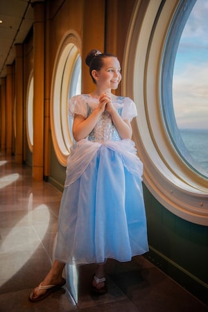 A young girl in a princess costume smiles while looking out a porthole window on a Disney cruise ship