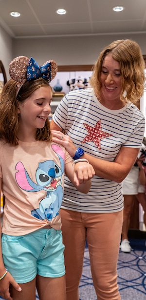 A woman smiling next to young girl wearing Minnie Mouse ears and a Stitch and Lilo tee shirt while looking down at the Disney Band Plus on her wrist