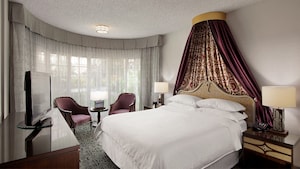 A bedroom with a bed, two chairs, lamps, a dresser and a TV