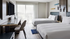 A hotel room features modern furniture that includes 2 double beds, a console, desk, lamps, chairs, an area rug and a flat screen television