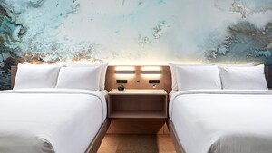 Two standard queen beds, a nightstand with a clock and wall-mounted lights between them 