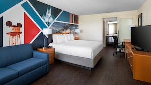 The interior of a room with a sofa, nightstands, king size bed, desk and chair, bureau, flat screen TV and Disney theme park inspired graphics on one wall