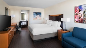 A sofa, nightstand and one king bed with headboard face a television on a dresser and desk with chair and beyond, a vanity area