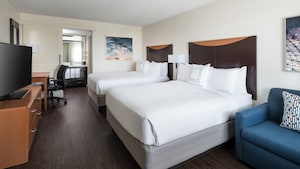 A sofa and 2 queen beds with headboards face a television on a dresser and desk with chair and beyond, a vanity area