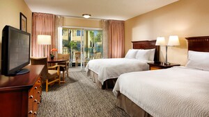 Two queen beds across from a TV-dresser, round table with 2 chairs and, beyond, a balcony