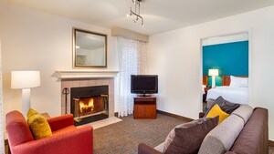The interior of a condo suite features a living area with a fireplace, TV, a small sofa, an upholstered chair and a doorway opening to a separate bedroom