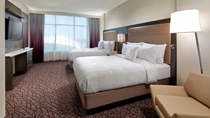 Bedroom has chair and lamp, 2 queen beds with headboards and shared nightstand facing a dresser and wall mounted television