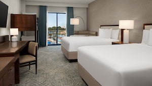 A hotel bedroom with 2 queen size beds, a bunk bed, nightstand, 2 lamps, a desk with chair, TV, a sofa, and a window with a view of Surfside Waterpark