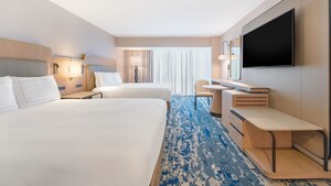 A hotel bedroom with a nightstand, desk, chair, table, mirror, TV and 2 queen beds