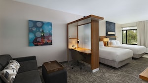 A small sitting area with a couch, coffee table and painting, and a desk that separates it from the bedroom area, with 2 queen beds