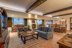 A common area with a sofa, 2 armchairs, a low table, an entertainment center with TV, a bench, a fireplace, a dining table and 2 windows with resort views in the Coco Suite at Pixar Place Hotel