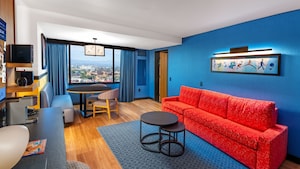 Un área de estar de una habitación de hotel con un comedor para cuatro personas, una lámpara Luxo Junior, un sofá, dos sillones orejeros, una mesa de centro, arte de Pixar en las paredes y puertas de vidrio que llevan a un balcón