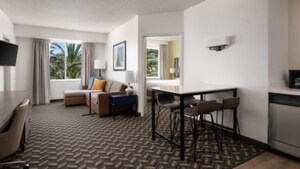 The Kids' Suite with a living area that includes an L-shaped couch, side tables, a TV, a desk and chair, a dining area that includes a high top table and 4 chairs, a small kitchen area, and a bedroom.