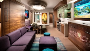 A hotel villa living area with a kitchen, couch, dining table, TV and 'The Princess and the Frog' themed artwork