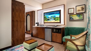 A hotel villa studio with 2 chairs, a TV, mini fridge, microwave, mirror and 'The Jungle Book'-themed artwork
