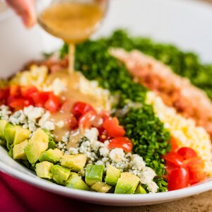 Salada cobb com abacate, bacon, alface, tomate e mais com molho de salada sendo servido por uma mão acima do prato