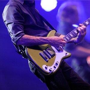 Um guitarrista se apresenta no palco em primeiro plano junto a outro músico tocando baixo