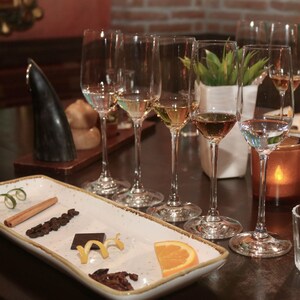 Uma mesa posta para degustação de tequila, com 5 taças, 2 copinhos de doses e um prato com casca de limão, canela em rama, grãos de café, um bloco de chocolate, casca de limão siciliano, gomo de laranja e gafanhotos fritos