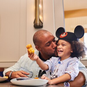 Homem beijando uma criança na bochecha, sentada em uma mesa com suco de laranja e waffles do Mickey