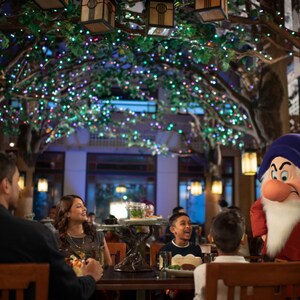 O anão Grumpy ao lado de uma mesa no Artist Point, com uma família de 4 pessoas fazendo uma refeição e sorrindo