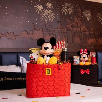 A Mickey Mouse plush in a gift box sit on a hotel bed