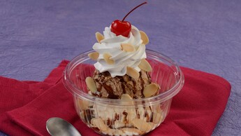 Dos bolas de helado en un vaso de plástico con ingredientes como crema batida, frutos secos y una cereza