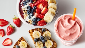 Fresas, un tazón de fruta en rodajas con granola y moras, un batido en una taza y una rebanada de pan francés cubierto con moras azules, bananas en rodajas y miel