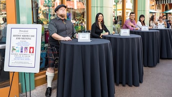 Hombres y mujeres están sentados en mesas redondas altas separadas y un letrero dice Disneyland Artist Showcase and Signing