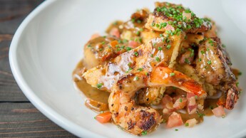 Un plato de camarones vudú con camarones asados, triángulos de pan de maíz, y tomates y cebollas picados en cubos en una salsa reducida, espolvoreado con cebollines picados.