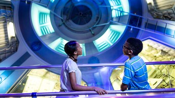 Un hermano y una hermana observan una rueda giratoria que simula la gravedad en el espacio infinito en la sección Mission: SPACE