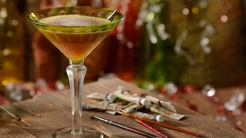 Un elegante vaso de martini lleno con un cóctel Night Monkey en una mesa al lado de tubos de acuarelas y pinceles