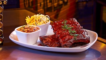 A rack of ribs covered with barbecue sauce on a plate with separate cups of baked beans and coleslaw
