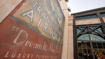 Un póster pintado a mano con tema de aviación frente a Maria and Enzo’s Ristorante con el texto “Trans Global Airways Dream It, Then Do It Luxury Flights Worldwide”