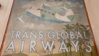 Un póster pintado a mano con tema de aviación frente a Maria and Enzo’s Ristorante con el texto Trans Global Airways
