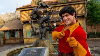 Gaston Character smiles and points in front of the fountain by Gastons Tavern
