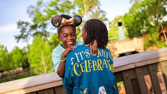 Criança sorridente usando uma tiara com orelhas da atração Tiana’s Bayou Adventure e abraçando uma mulher que está usando uma jaqueta da atração Tiana’s Bayou Adventure na qual se lê: “It’s a joyful celebration”
