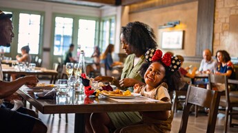 A young girl wearing Minnie Mouse ears sits at a table in a restaurant eating with her mother